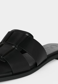 Μαύρο σανδάλι που φοριέται εύκολα, με φαρδιές δερμάτινες λωρίδες που διασταυρώνονται πάνω στο πόδι και επίπεδη σόλα, εμφανίζεται σε απλό λευκό φόντο.