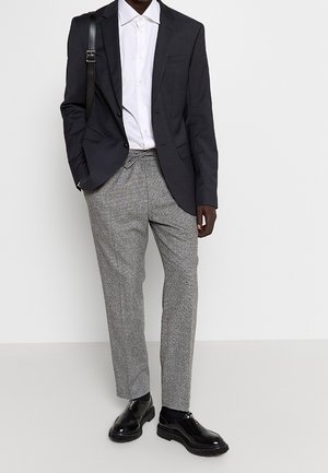 Homme portant un blazer noir, une chemise blanche avec des boutons, un pantalon gris à cordon, des chaussures de ville noires et un sac bandoulière noir sur fond blanc.