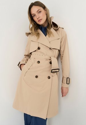 Kvinde iført beige dobbeltbrystet trenchcoat med bælte og spænde detaljer, stående mod en hvid væg.