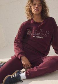 Maroonsweater og bukser sæt med teksten "S.E.L.F. WELLNESS CLUB". Model iført marineblå sneakers og hvide sokker, siddende på en lys overflade.
