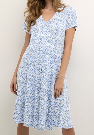 Kvinde iført en knælængde kjole med korte ærmer, med et lille blåt og hvidt blomsterprint og v-hals.