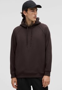 Brauner Hoodie aus einem weichen Material, mit einer Kapuze mit Kordelzug, Raglanärmel und elastischem Saum. Trägt eine schwarze Mütze.