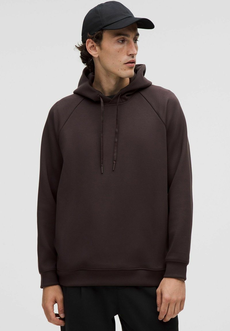 Brauner Hoodie aus einem weichen Material, mit einer Kapuze mit Kordelzug, Raglanärmel und elastischem Saum. Trägt eine schwarze Mütze.