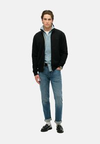 Felpa nera con zip sopra una camicia di denim azzurra chiara, abbinata a jeans azzurri chiari con risvolto e scarpe nere. Il modello è in piedi contro uno sfondo bianco.