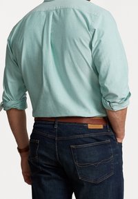 Uomo con camicia a maniche lunghe verde chiaro con le maniche arrotolate, infilata in jeans blu scuro e cintura in pelle marrone, mani in tasca.