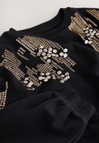 Črna jopa z zlatimi bleščicami v vzorcu valov, z rebrastim manšetnim detajlom. Mehka tekstura tkanine z običajnim krojem.