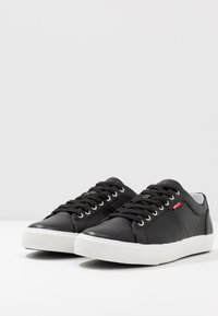 Fekete bőr sneaker cipők sima felületűek, fehér gumitalppal, fekete fűzőkkel és apró piros logó részlettel az oldalukon.