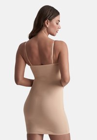 Vrouw met lang bruin haar in een strakke naadloze slipdress met dunne bandjes, van achteren gezien tegen een effen achtergrond.