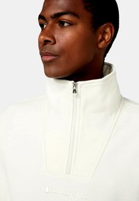 Sudadera de forro polar blanco con media cremallera y cuello alto, presenta un pequeño logo en la parte inferior y detalles de costura visibles. Textura suave.