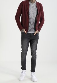Chaqueta de cremallera color marrón sobre una camiseta gris con estampado, combinada con jeans negros ajustados y zapatillas blancas. Textura suave, corte casual.
