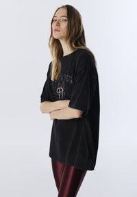 Czarna, oversize koszulka z grafiką i perłowymi detalami z przodu, w połączeniu z błyszczącymi bordowymi legginsami. Gładka faktura materiału.