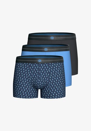 Oberkörperfreier Mann steht, trägt blaue Boxershorts mit schwarzem Bund, vor einem schlichten hellen Hintergrund.