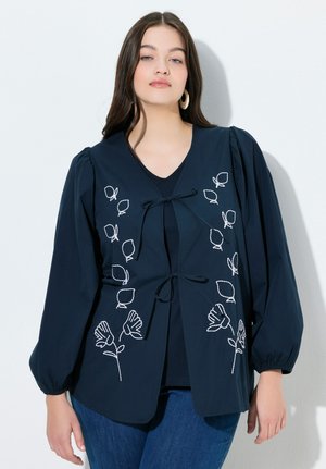 Kvinde iført en mørkeblå bluse med puffede lange ærmer og hvid blomsterbroderi, kombineret med blå jeans, stående mod en hvid væg.