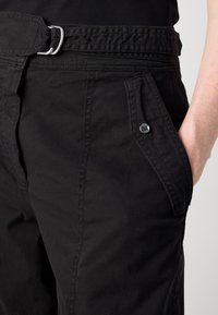 Primo piano di pantaloni neri con una patta della tasca cucita fissata da un bottone a pressione metallico e una cintura in tessuto nera abbinata con fibbia argentata.