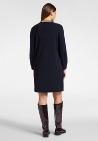 Frau von hinten, die ein knielanges, marineblaues Kleid mit langen Ärmeln und dunkelbraunen kniehohen Stiefeln trägt, vor einem schlichten Hintergrund.