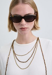 Gafas de sol con una montura gruesa de carey y lentes oscuras, combinadas con tres collares metálicos dorados en capas, usados sobre una camisa blanca.