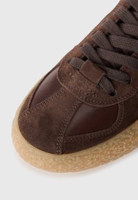 Sneaker en daim et cuir marron avec une semelle en caoutchouc texturée, dotée de lacets marron et de coutures ton sur ton. Design à bout rond.