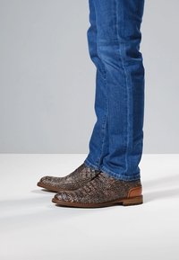 Scarpe marroni con texture a motivo rettiliano, caratterizzate da un tacco basso e dettagli marroni, abbinate a jeans di denim blu.