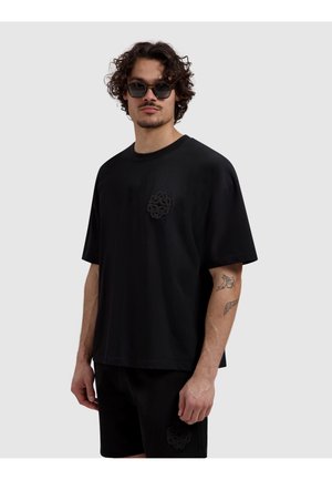 Man with curly hair and sunglasses wearing black oversized t-shirt and shorts with black embroidered logos on chest and leg.