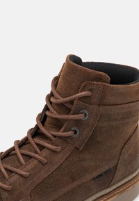 Botine înalte din suede maro cu finisaj texturat, vârf rotund, ochiuri metalice și sireturi maro în contrast. Prezintă detalii pe latură cu marcaj și guler căptușit.