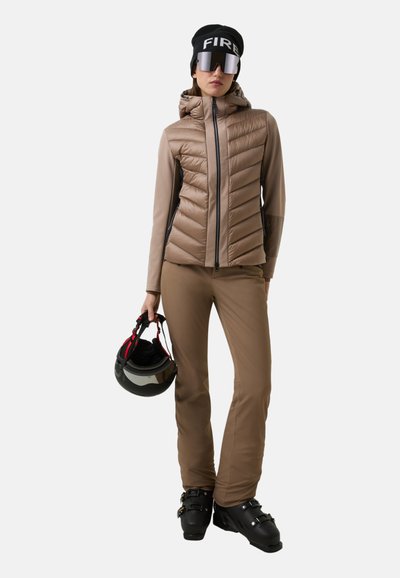Kobieta w brązowym ubraniu narciarskim trzyma czarny kask, ma na sobie czarne buty narciarskie, lustrzane gogle i czapkę beanie w kolorze czarnym z napisem "FIRE".