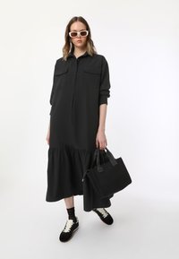 Robe noire boutonnée avec un ourlet à volants en couches, deux poches poitrine, associée à un sac à main noir et des baskets noires avec des lacets blancs.