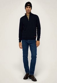 Marinblå tröja med dragkedja i halsen, gjord av stickat material, bärs med blå jeans. Outfitten inkluderar en svart mössa och bruna skor med slät yta.