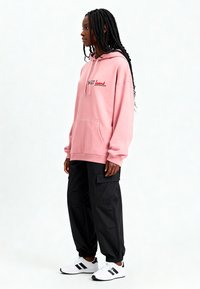 Hoodie rosa con il testo "sweet heart", abbinato a pantaloni cargo neri e sneakers bianche. Caratterizzato da una tasca a marsupio e una vestibilità rilassata.