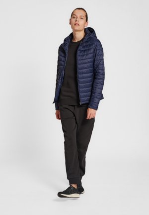 Marineblauwe gewatteerde jas met capuchon, met een quilted ontwerp, ritssluiting aan de voorkant en zijzakken, gecombineerd met een zwarte sweatshirt en broek.