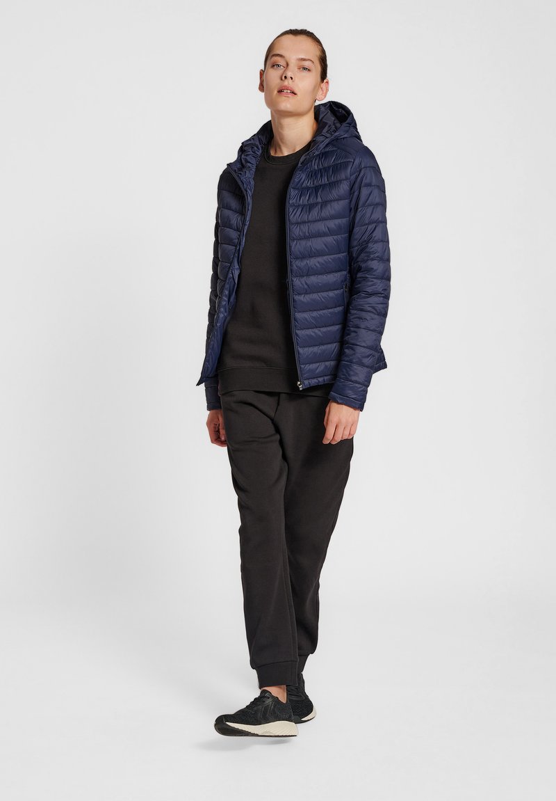 Marineblauwe gewatteerde jas met capuchon, met een quilted ontwerp, ritssluiting aan de voorkant en zijzakken, gecombineerd met een zwarte sweatshirt en broek.