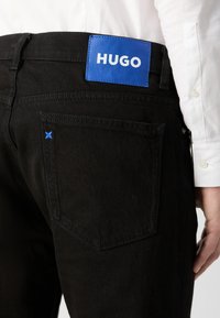 Jeans en denim noir avec une étiquette en cuir bleue portant le logo « HUGO ». Détails de couture visibles et accent bleu sur la poche arrière.