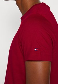Rotes T-Shirt mit kurzen Ärmeln aus Baumwolle, mit einem kleinen gestickten Logo in Blau, Weiß und Rot auf dem linken Ärmel. Regelmäßiger Schnitt.
