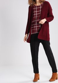 Bordeauxfarbener Strickcardigan mit strukturiertem Design, kombiniert mit einem bordeaux- und weißkarierten Oberteil und schwarzen, tapered Hosen, abgerundet mit braunen Ankle Boots.