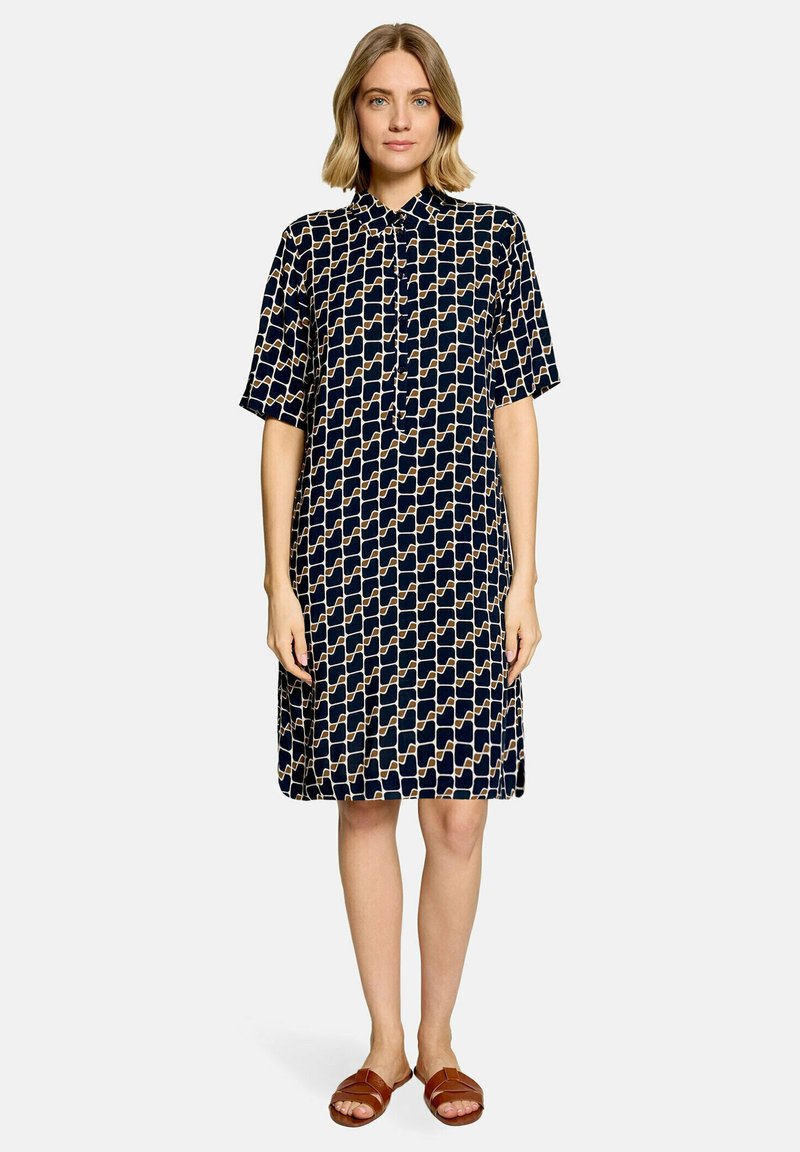 Femme aux cheveux blonds courts portant une robe marine à manches courtes avec un motif géométrique beige et des sandales plates marron, debout sur un fond blanc.