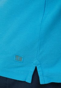 Turkusowa bawełniana koszulka polo o teksturowanej powierzchni, z małą metką marki na dole i bocznym rozporkiem u dołu.