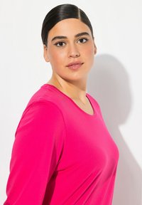 Enganliegende Langarmbluse in leuchtendem Pink, aus glattem Stoff mit Rundhalsausschnitt und dezentem Glanz. Ohne Muster oder Verzierungen.