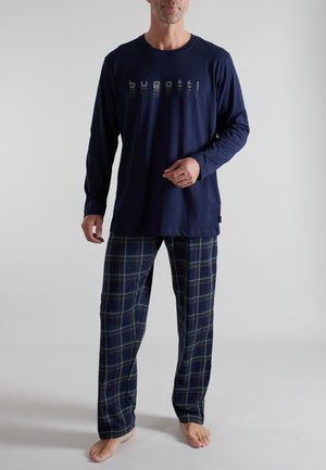 Mann trägt navyblaues Langarmshirt mit "bugatti"-Schriftzug und blaue karierte Schlafanzughose, steht barfuß vor einem einfarbigen Hintergrund.