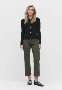 Musta ribbed neulejakku pyöreällä kaula-aukolla ja nappikiinnityksellä, yhdistettynä oliivinvihreisiin räätälöityihin housuihin ja mustiin liukuviin kenkiin.