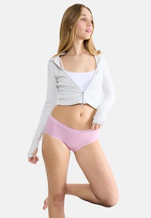 Woman wearing a white cropped zip-up top over a white tank and light pink underwear, posing with one leg bent against a white background.