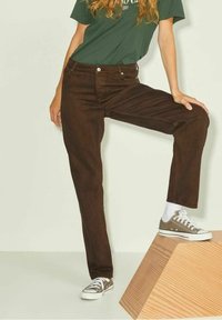 Jeans de mezclilla marrón con un corte recto, bolsillos frontales y acentos de botón de metal. Combinados con una camiseta verde oscura y zapatillas grises.