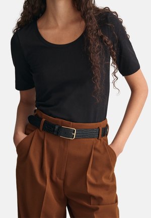 Personne portant une chemise noire à manches courtes rentrée dans un pantalon marron taille haute avec une ceinture noire texturée et les mains dans les poches.