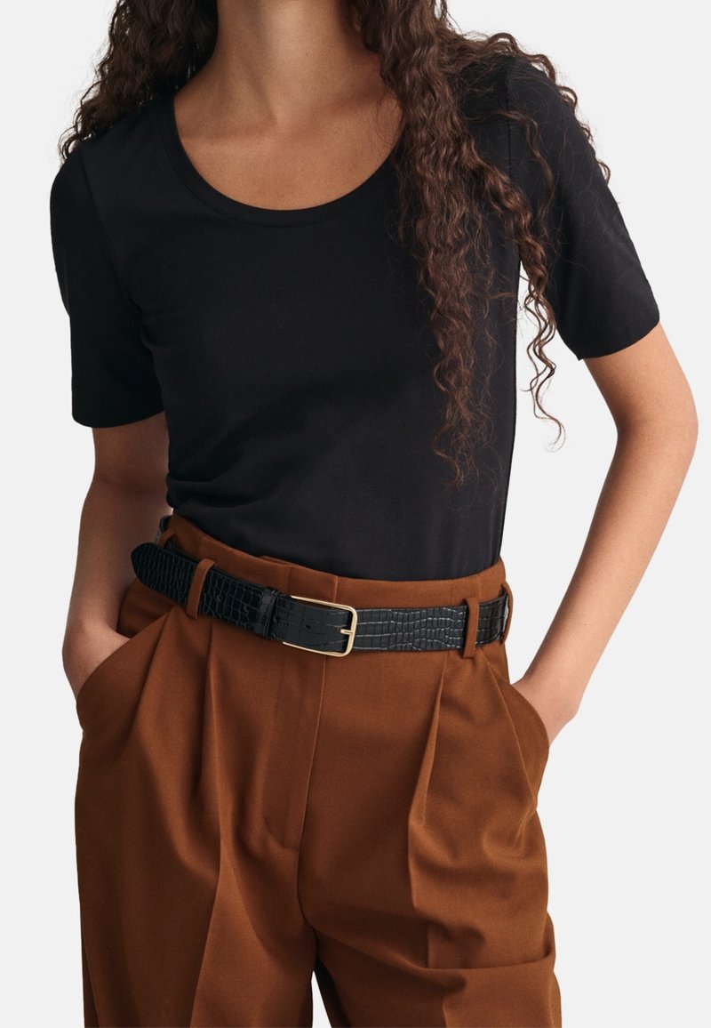 Personne portant une chemise noire à manches courtes rentrée dans un pantalon marron taille haute avec une ceinture noire texturée et les mains dans les poches.