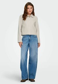 Donna in piedi con una giacca beige abbottonata, jeans blu a gamba larga e scarpe marroni su uno sfondo chiaro e semplice.