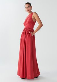 Abito lungo rosso senza maniche con bustino aderente, vita arricciata e tasche laterali. Tessuto morbido con gonna fluente, design minimale.