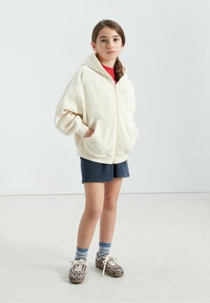 Hoodie zippé en polaire crème avec poches avant, porté sur un t-shirt rouge, associé à un short bleu marine, des chaussures à imprimé léopard et des chaussettes rayées.