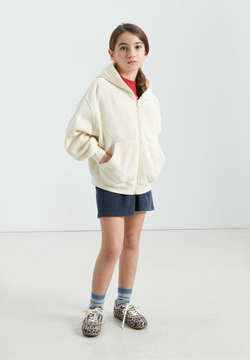 Hoodie zippé en polaire crème avec poches avant, porté sur un t-shirt rouge, associé à un short bleu marine, des chaussures à imprimé léopard et des chaussettes rayées.