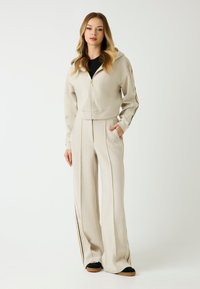 Beżowa bluza z zamkiem błyskawicznym oraz pasujące spodnie dresowe, wykonane z miękkiego materiału, z czarnymi bocznymi paskami i przednimi kieszeniami.