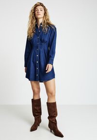 Denim-Hemdkleid mit tailliertem Schnitt, langen Ärmeln und durchgehendem Knopfverschluss. Mit goldenen Knöpfen und einem leicht ausgestellten Rock. Braune knielange Stiefel.