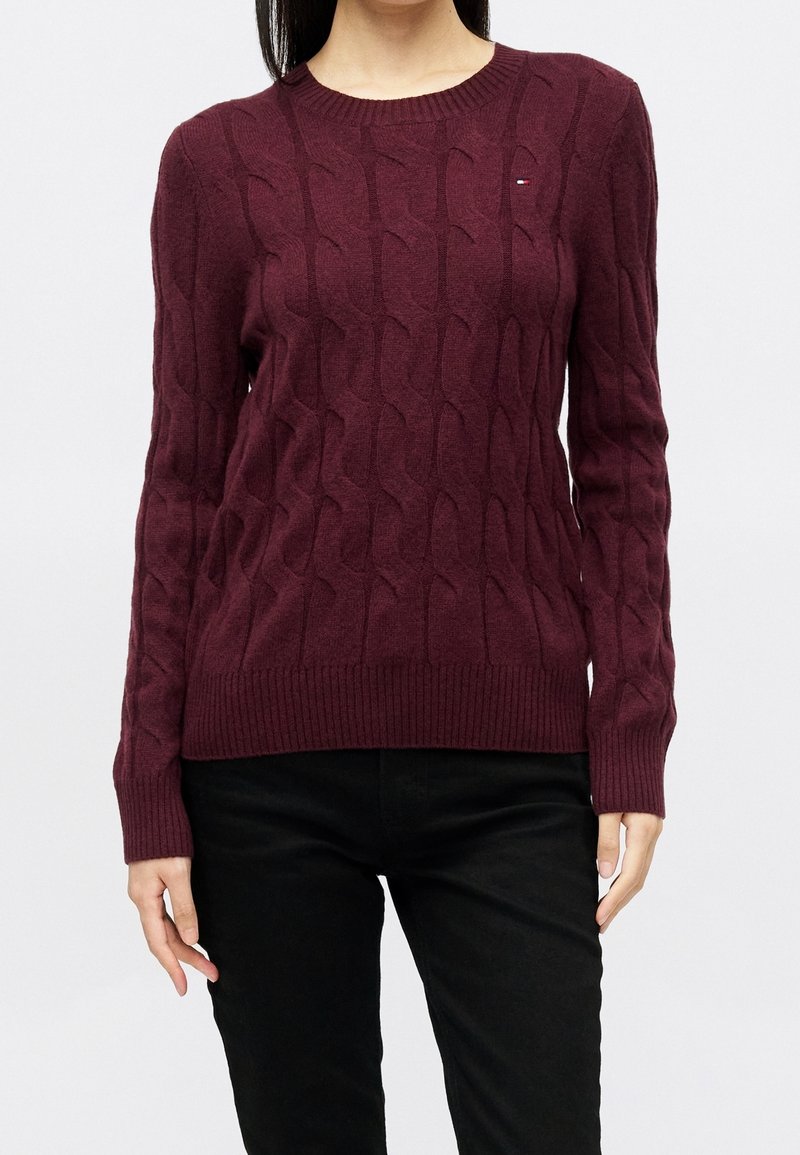 Femme portant un pull torsadé bordeaux à manches longues et un pantalon noir, debout devant un fond clair uni.