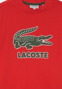 Camiseta de algodón roja con un logo de cocodrilo verde y el texto "LACOSTE" en verde, contorneado en blanco. Diseño de manga corta y cuello redondo.